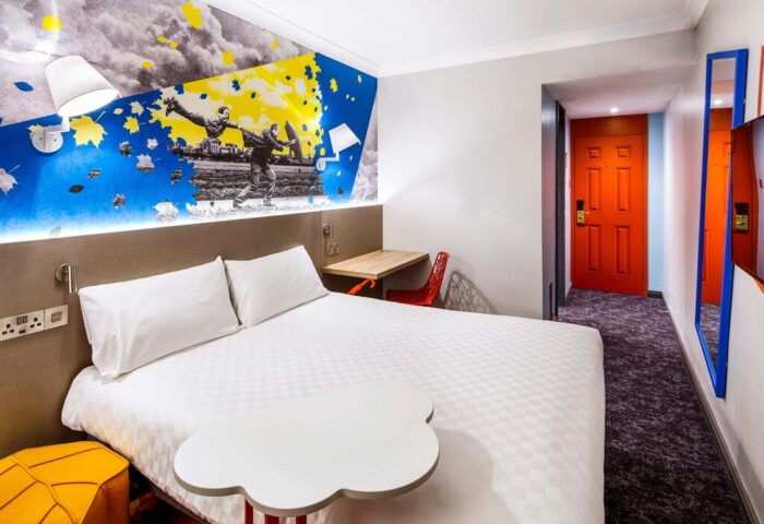 Ibis Styles Manchester Portland gay-friendly hotel room. Bed, desk, orange door, & colorful wall art. Modern design for gay travelers.