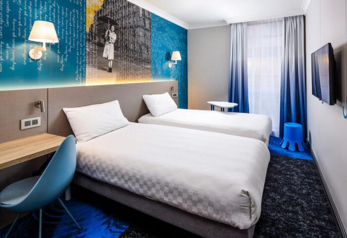 ibis Styles Manchester Portland hotel room. Two twin beds with blue themed decor. Gay-friendly hotel for gay men travel advice.