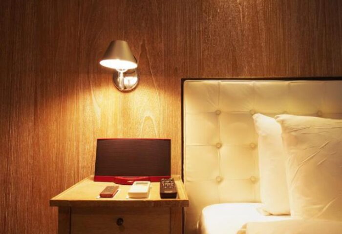 Inhouse Hotel detail: Bedside table with remotes, speaker, and lamp. Gay-friendly hotel room with wood paneling and tufted headboard.