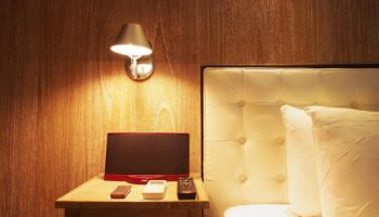 Inhouse Hotel detail: Bedside table with remotes, speaker, and lamp. Gay-friendly hotel room with wood paneling and tufted headboard.