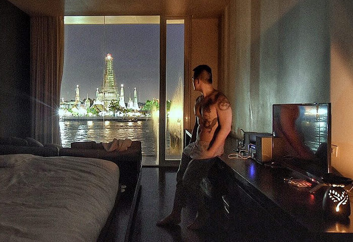 Gay-friendly Sala Rattanakosin Bangkok hotel room. Man with tattoos looks out at Wat Arun temple at night. Travel advice for gay men.