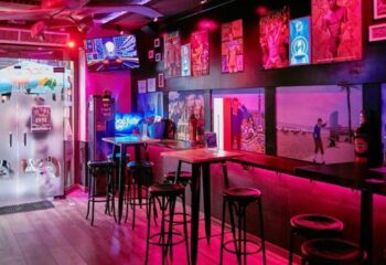 GinGin Gay Bar in Barcelona: Interior view of the bar with pink lighting, stools, tables, and wall art. Popular gay travel destination.