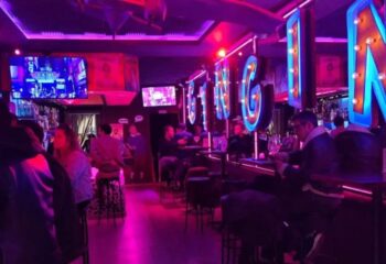 Gin Gin Gay Bar, Barcelona. Interior view of the bar with patrons seated at the bar and tables, lit with neon lights. Gay travel.