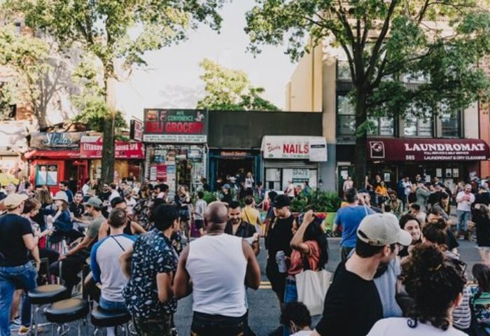 Good Judy Bar for queers in New York. Outdoor crowd scene, people gathered on the street in front of shops, including a laundromat.