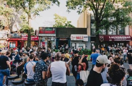 Good Judy Bar for queers in New York. Outdoor crowd scene, people gathered on the street in front of shops, including a laundromat.