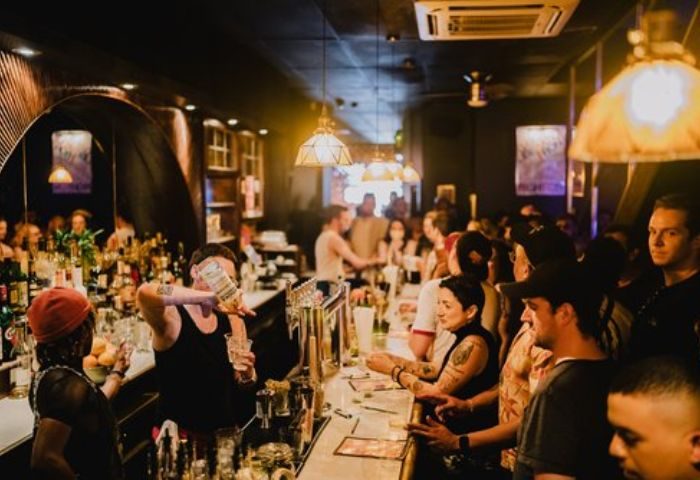 Good Judy Bar, New York. Bartender pouring drinks for a crowded bar. Gay bar scene with people enjoying drinks. Queer nightlife.