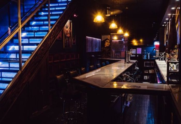 Good Judy Bar for queers in New York. Interior view of the bar with blue-lit stairs, bar counter, and seating. A gay travel destination.