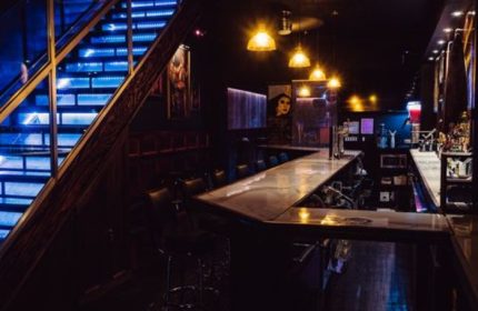 Good Judy Bar for queers in New York. Interior view of the bar with blue-lit stairs, bar counter, and seating. A gay travel destination.