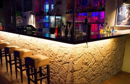 Grande Opera Frankfurt: Bar interior with stone facade, bar stools, illuminated bottles. Gay travel destination, vibrant nightlife.