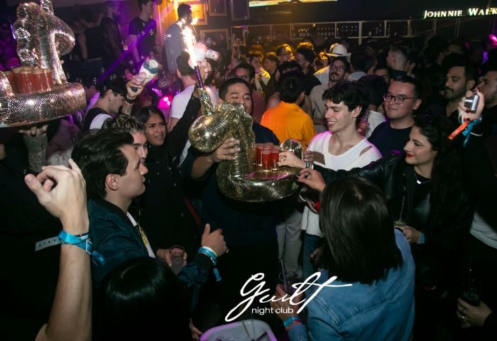 Guilt Night Club, a gay bar in Mexico. A crowd of people are gathered, some holding drinks on a unicorn-shaped tray. Nightlife scene.
