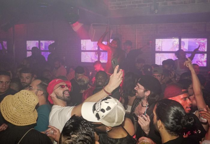 Inside HUSH HK gay bar, New York. A male dancer performs on a pole amidst a lively crowd in a dance club setting. Nightlife scene.