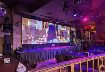 Hamburger Mary's Long Beach front stage view. Drag show venue with a large screen, stage lighting, and tables for dining. Gay travel.