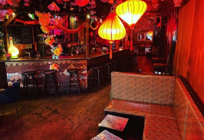 Interior of Happyfun Hideaway gay bar in New York. Bar stools line the bar. Lanterns hang from the ceiling. Booth seating is available.
