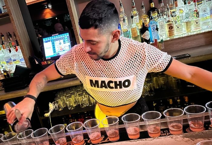 Bartender at Hardware Bar in New York. Man in MACHO shirt pours drinks. Gay nightlife, travel advice, bar scene. Cocktails.