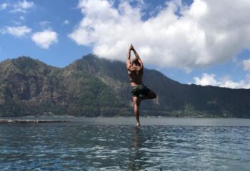 Gay man doing yoga by a lake with mountains. A shirtless man is in tree pose, hands together, in a pool. Serene gay travel.