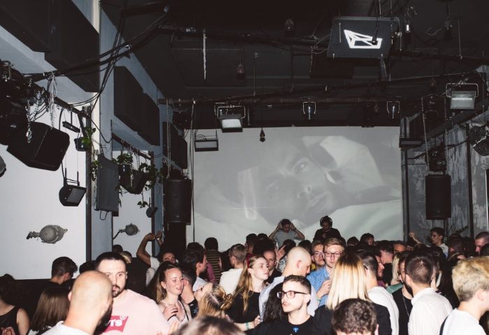 Hello Party at Toldi Club, Budapest. Crowd of people dancing, DJ on stage, screen displaying a close-up of a face. Gay nightlife.