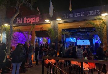 Hi Tops gay bar location. Exterior view of the Hi-Tops bar with a crowd of people. Cold pitchers hot catchers sign. Gay travel advice.