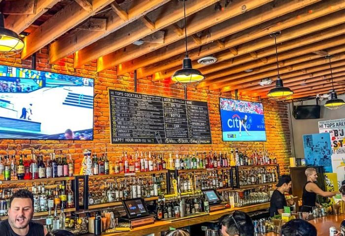 Hi Tops bar interior, morning. Brick walls, wooden ceiling, TVs showing sports. Menu of cocktails and drinks. Gay bar, travel advice.