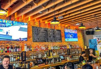 Hi Tops bar interior, morning. Brick walls, wooden ceiling, TVs showing sports. Menu of cocktails and drinks. Gay bar, travel advice.
