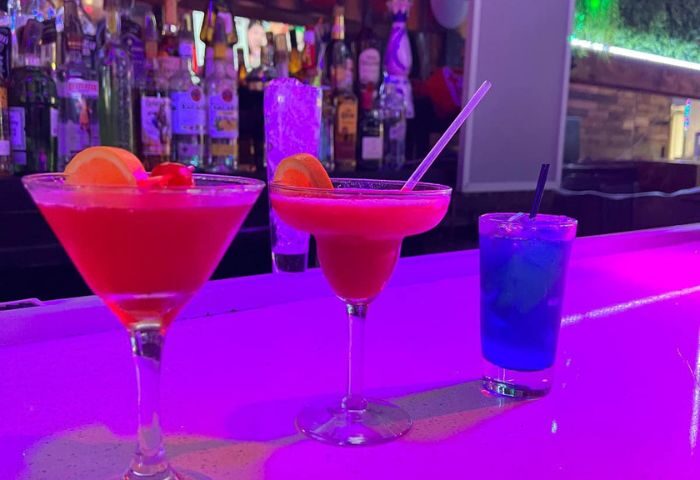 Cocktails at Hombres Lounge, New York. Three colorful drinks on a bar, lit with purple light. Gay travel advice for nightlife.