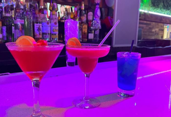 Cocktails at Hombres Lounge, New York. Three colorful drinks on a bar, lit with purple light. Gay travel advice for nightlife.