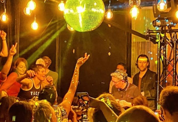 Homoelectric Manchester club scene. Gay men dancing under disco ball, lights. DJ playing music. Gay travel advice.