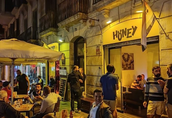 Honey furry gay bar in Barcelona. Exterior view of the bar at night with people sitting at tables and standing outside. Gay travel advice.