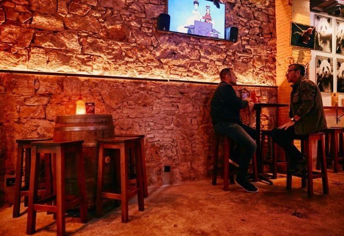 Honey Furry Gay Bar in Barcelona: Two men at a small table in a bar with stone walls, wooden stools, and a TV showing cartoons.