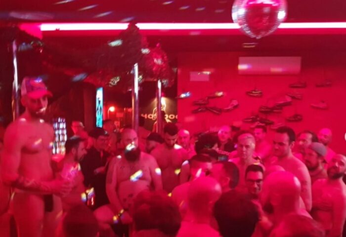 Inside Hot Dog Club Milan, a gay sauna, men gather under red lights & disco ball. Some are shirtless, enjoying the social atmosphere.
