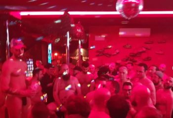 Inside Hot Dog Club Milan, a gay sauna, men gather under red lights & disco ball. Some are shirtless, enjoying the social atmosphere.