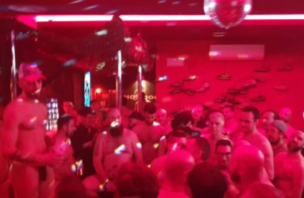 Inside Hot Dog Club Milan, a gay sauna, men gather under red lights & disco ball. Some are shirtless, enjoying the social atmosphere.
