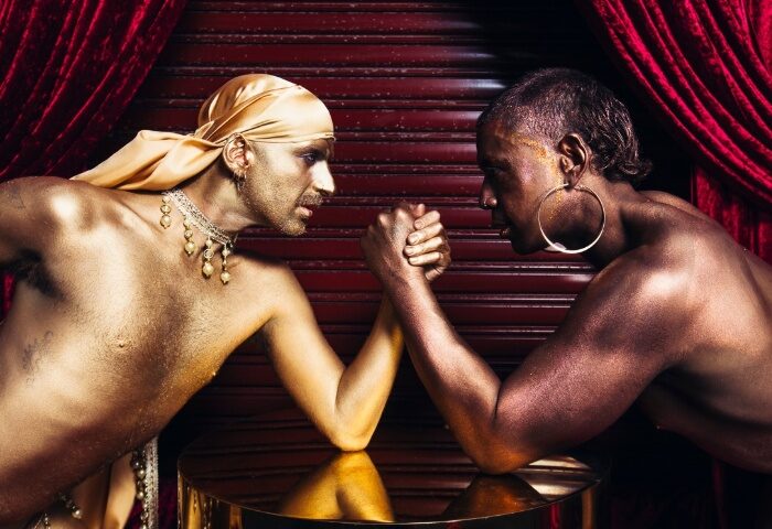 House of Mince Sydney: Two shirtless men arm wrestling, bodies painted gold and bronze. Red curtain backdrop. Gay Sydney travel.