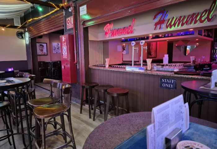 Interior of Hummel Hummel, a gay bar in Gran Canaria. Bar stools line the bar. Tables and chairs fill the space. Staff Only sign.