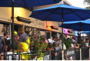 Outdoor view of Hunters Nightclub, Palm Springs. Gay bar scene with blue umbrellas, heaters, and a crowd of people. Popular gay travel destination.