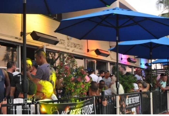 Outdoor view of Hunters Nightclub, Palm Springs. Gay bar scene with blue umbrellas, heaters, and a crowd of people. Popular gay travel destination.