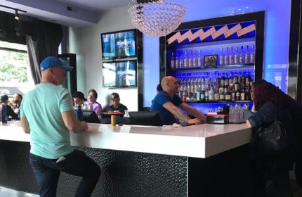 Inside Hydrate Nightclub, Chicago. Bartender serves patrons. TVs, chandelier, & liquor bottles. Gay bar scene, nightlife.