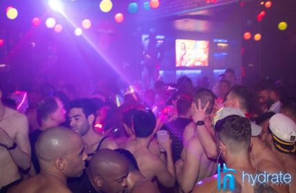 Crowded Hydrate gay bar in Chicago. Shirtless men dance under purple lights. A drag queen performs on stage. Nightlife scene.