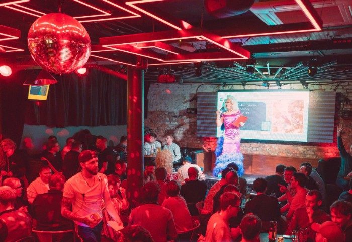 Inside Iconic Bar Manchester: Drag queen performance on stage for a crowd of gay men. Disco ball, red lighting, vibrant atmosphere.