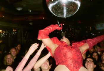 Imperial Erskineville Sydney: Drag queen in red performs under a disco ball, cheered on by a crowd. Gay travel nightlife scene.