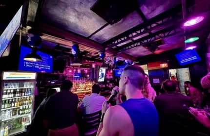 In Between Waikiki gay bar karaoke in Honolulu. A man sings at a bar with a rainbow flag and TV screens with karaoke lyrics.