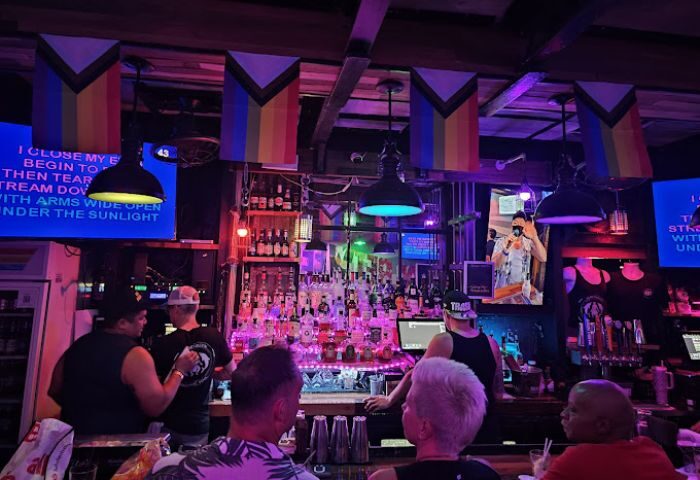 In Between Waikiki gay bar karaoke in Honolulu. Rainbow flags hang above a busy bar with patrons and a karaoke screen. Gay travel.