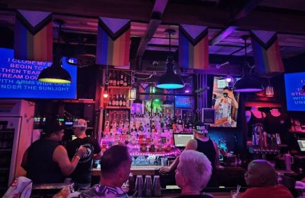 In Between Waikiki gay bar karaoke in Honolulu. Rainbow flags hang above a busy bar with patrons and a karaoke screen. Gay travel.