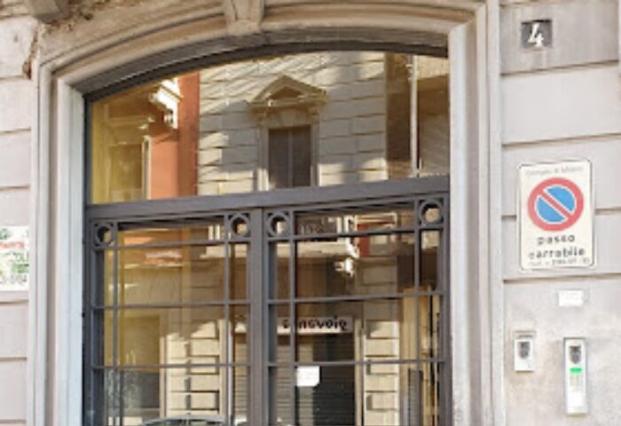 Inferus Club Milan entrance. Metal-framed glass doors reflect the building opposite. A no parking sign is visible on the wall. Gay travel.