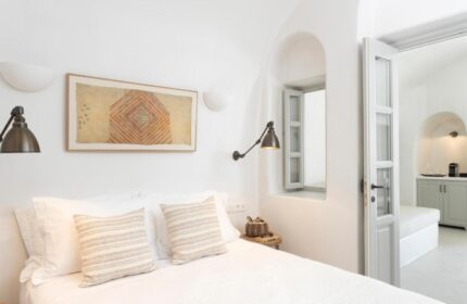 Armenaki Santorini hotel room. Interior of a luxury gay-friendly hotel with rooftop pool. White walls, bed with pillows, window, and door to another room.