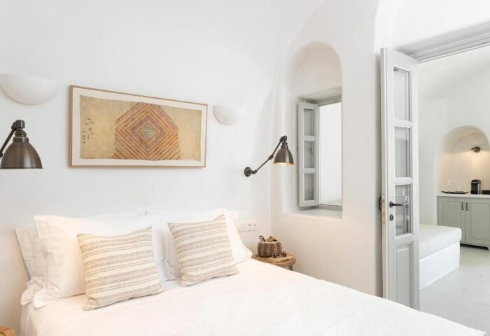 Armenaki Santorini hotel room. Interior of a luxury gay-friendly hotel with rooftop pool. White walls, bed with pillows, window, and door to another room.