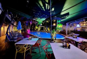 Island House Key West pool at night. Gay cruising resort with tables, chairs, bar, palm trees, and colorful lighting.