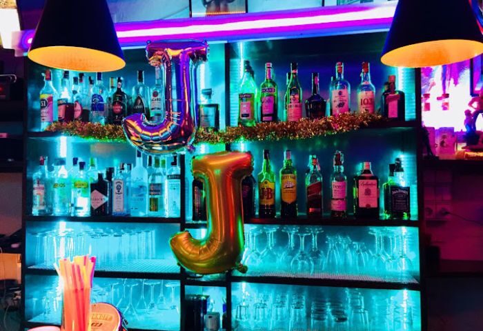 JJ Bar, a gay bar in Ibiza, Spain. Shelves are stocked with liquor bottles, glasses, and J-shaped balloons. Neon lights illuminate the scene.