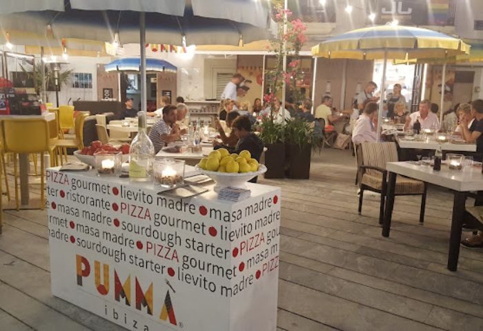 JJ Bar gay bar in Ibiza: outdoor seating at a restaurant with people dining under umbrellas. Pumma Ibiza stand with lemons/tomatoes visible.