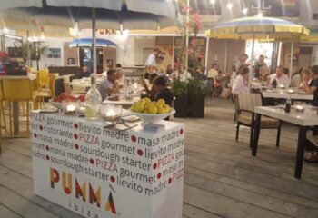 JJ Bar gay bar in Ibiza: outdoor seating at a restaurant with people dining under umbrellas. Pumma Ibiza stand with lemons/tomatoes visible.