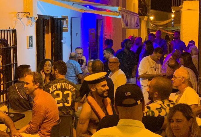 Ibiza gay bar scene at JJ Bar: Men enjoy drinks and socializing. A sailor-themed patron stands out in this vibrant gay nightlife spot.